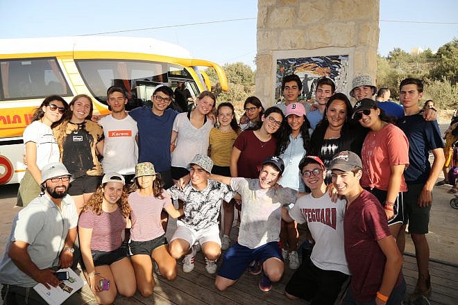 Ruth Schwartz (second from right, middle row), the mother of Ezra Schwartz, stands at the “Ezra Schwartz Lookout,” surrounded by teens in Israel from Camp Yavneh, where her son went to summer camp. Credit: Photo by Gershon Alanson.