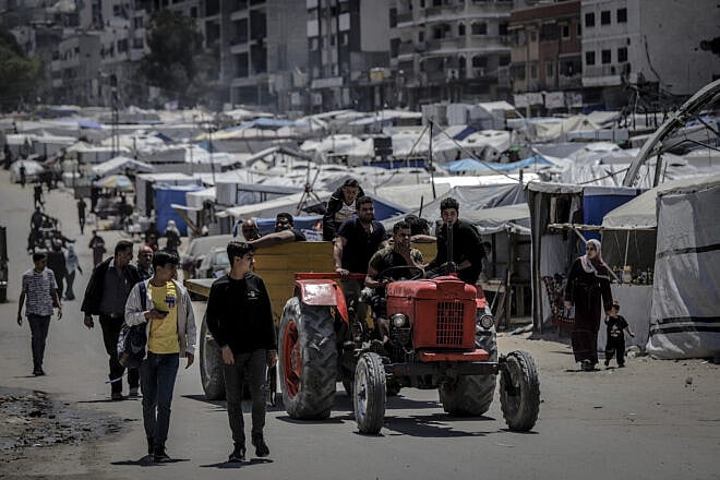 A man drives a red tractor past pedestrians and tents lining the street in the Al-Rimal neighborhood of Gaza City, on May 4, 2025. Photo by Saeed Jaras/Middle East Images/AFP via Getty Images.